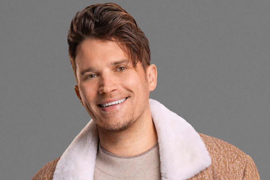 Tom Schwartz smiling in a beige sweater under a light brown and white fur trimmed coat and posing in front of a grey backdrop.