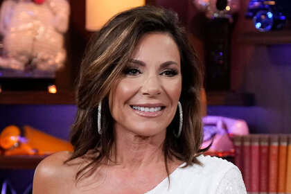 Luann smiling in a white, one shoulder, dress with hoop earrings.