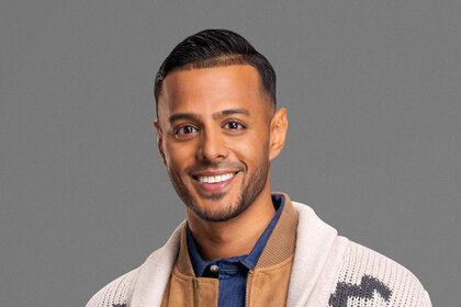 Brian Benni smiling and wearing a white patterned cardigan over a brown, suede, jacket and denim shirt in front of a grey backdrop.
