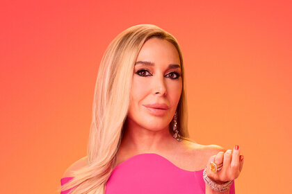 Marysol Patton posing in a long-sleeved, off shoulder, pink floor length gown in front of a red and orange background.