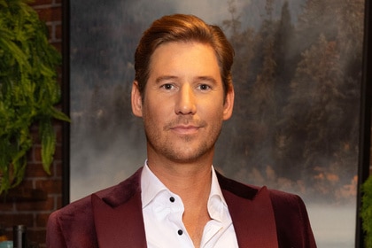 Austen Kroll wearing a velvet, maroon, suit jacket and white button down standing in front of a rustic seating area.