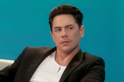 Tom Sandoval wearing a black blazer and a white tee shirt in front of a blue backdrop