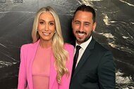 Heather Altman in a pink suit with a mesh top and Josh Altman in a black suit smile together in front of a black marble wall.