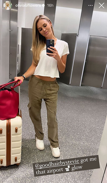 Olivia Flowers posing in a white t-shirt and olive pants with her luggage in an airport bathroom. Overlaid text: "@goodhairbystevie got me that airport [toilet emoji] glow".
