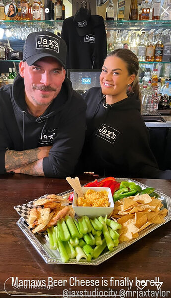 Jax Taylor and Brittany Cartwright of Vanderpump Rules smile in matching hoodies with a snack platter in front of them.