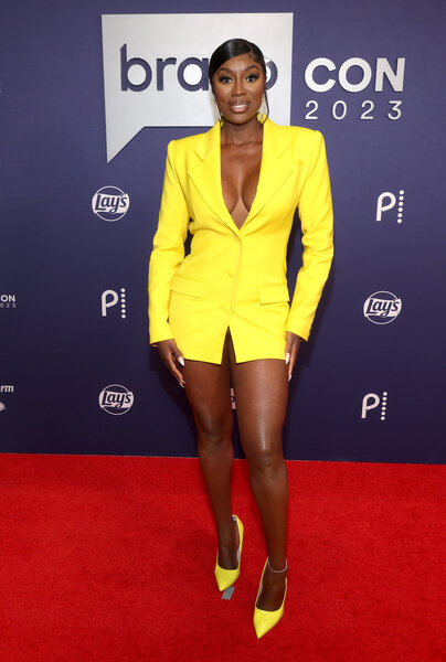 Wendy Osefo poses in a yellow dress and shoes on the red carpet of Bravocon 2023.
