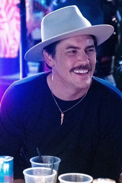Tom Sandoval smiling in a hat, black shirt, and lightning bolt necklace.