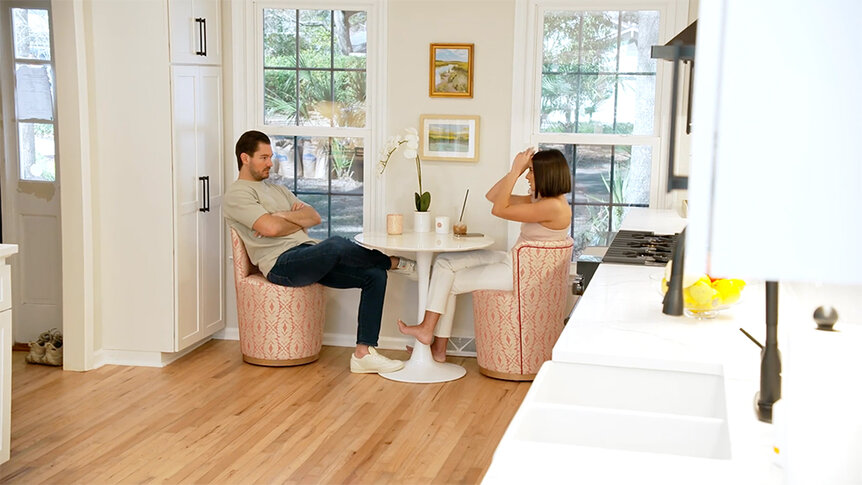 Craig Conover and Paige DeSorbo sitting and talking together at a round white table by two windows in Craig’s kitchen.