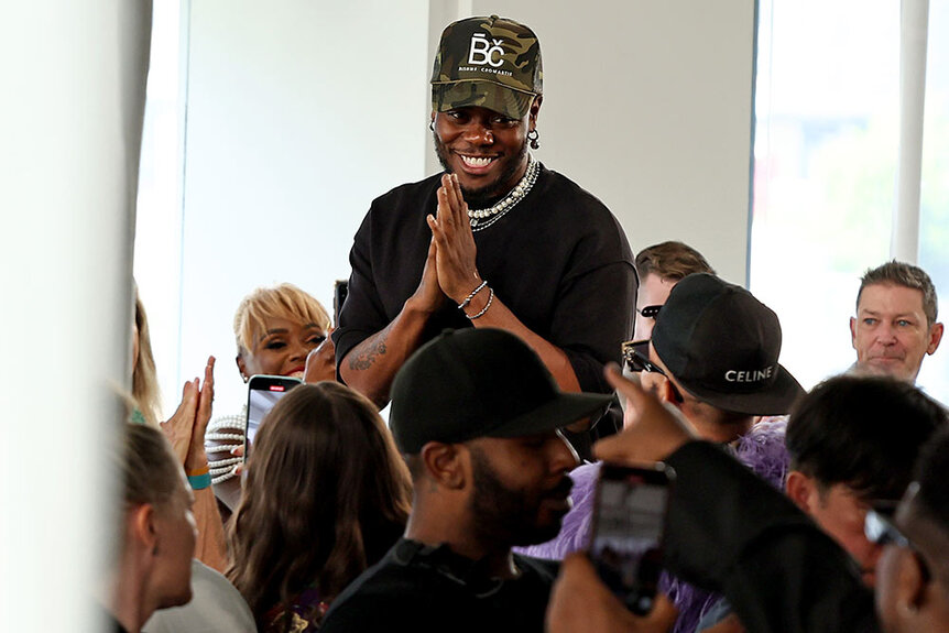 Bishme Cromartie walking on the runway at his show thanking his attendees wearing a camo baseball cap and black shirt.