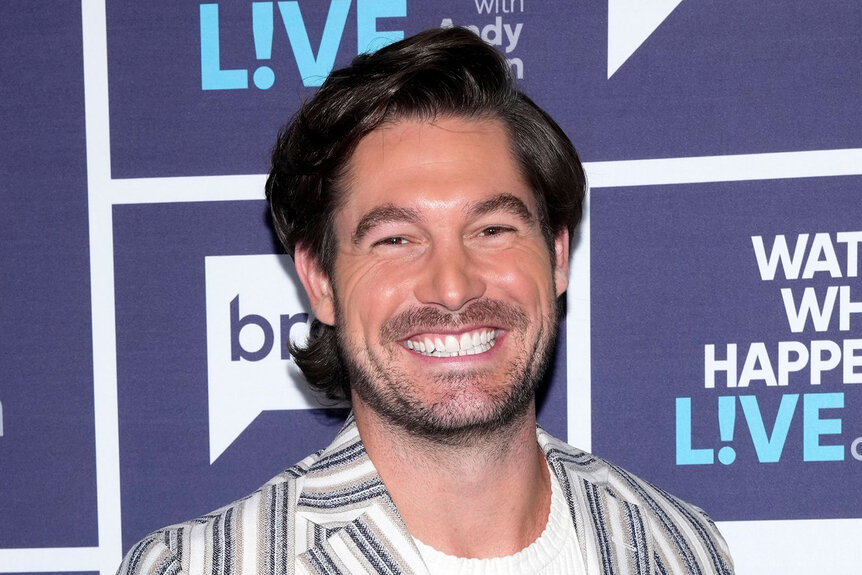 Craig Conover smiling in a striped black and white suit in front of the WWHL step and repeat.