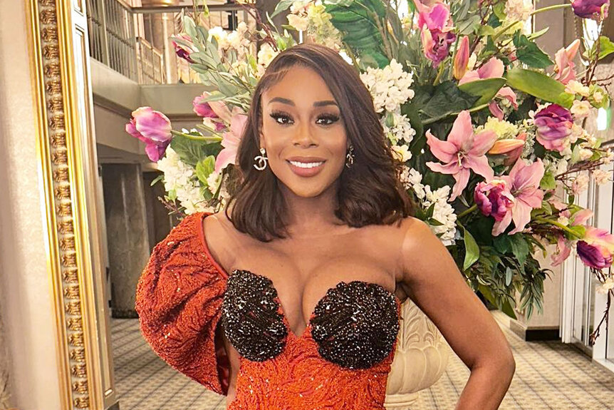 Nneka Ihim posing in an orange, one shoulder, gown in front of a large flower bouquet.