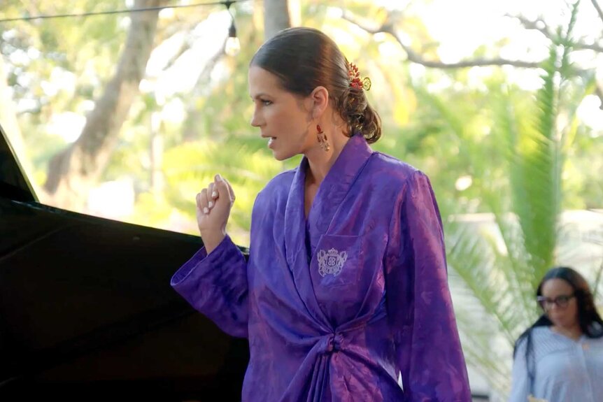 Julia, wearing a purple robe, is singing next to a piano.