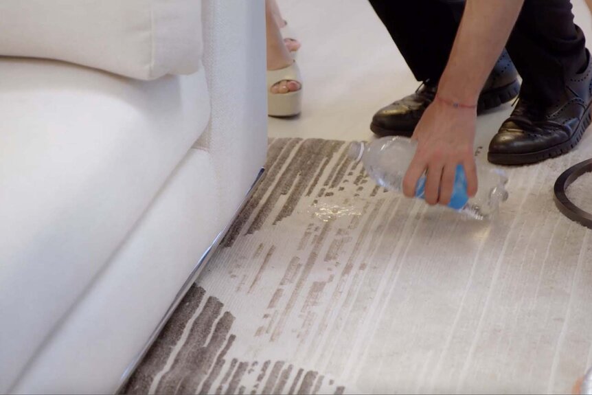 A hand pours a water bottle over a stain on a beige rug.