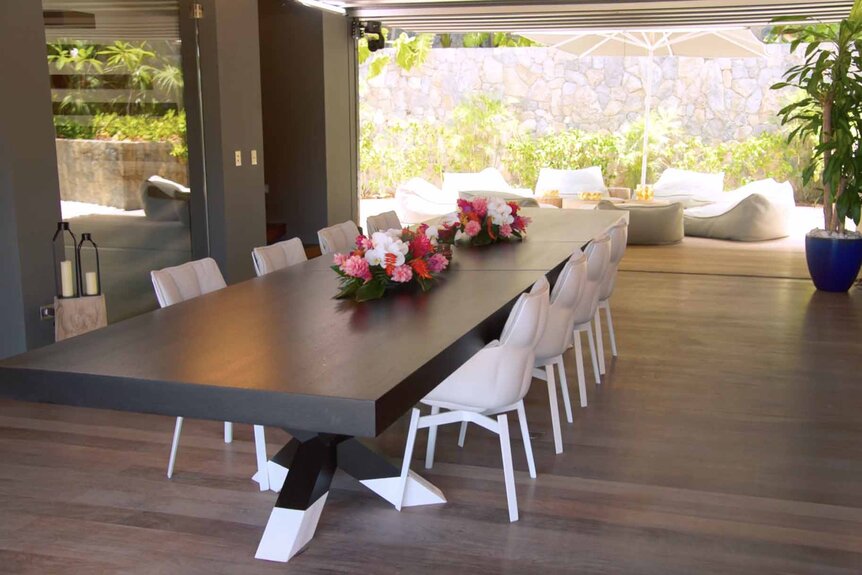 A long dining table with floral arrangements on top and an opening to white porch furniture.