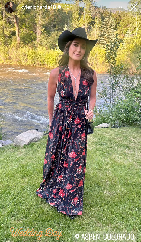 Kyle Richards poses in a black, floral, maxi dress and a black hat in front of a river.