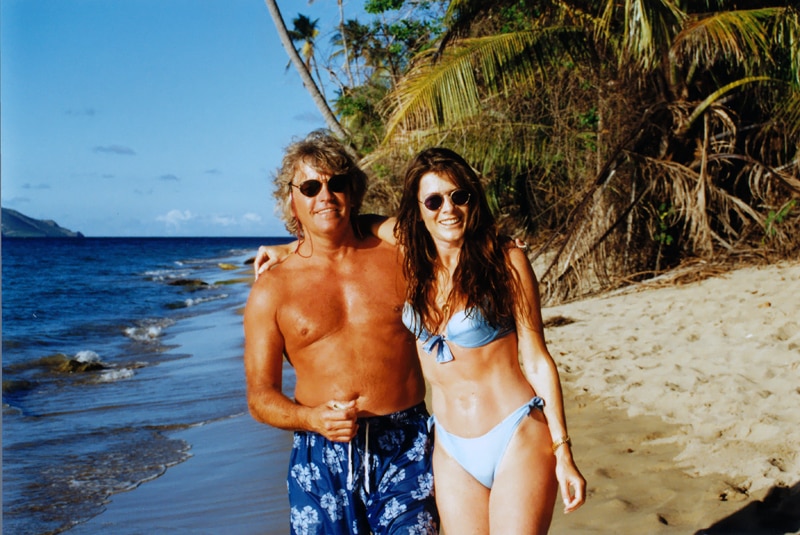 Ken and Lisa wearing blue swimwear and sunglasses while posing on a scenic beach together.