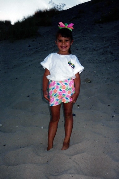Scheana Shay wears a colorful pair of shorts and bow while standing on the beach as a young kid.
