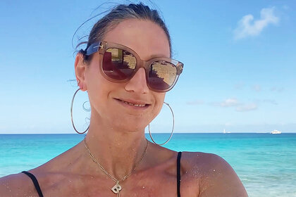 Jenna Lyons smiling with sunglasses and a gold necklace in front of the ocean.