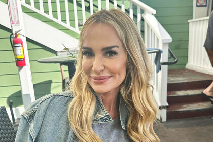 Taylor Armstrong smiling in a gray sweater and denim jacket in front of a green building with white trim.