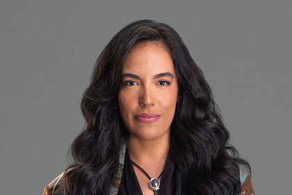 Danielle Olivera wearing a patterned coat, black shirt, and necklace in front of a grey backdrop.