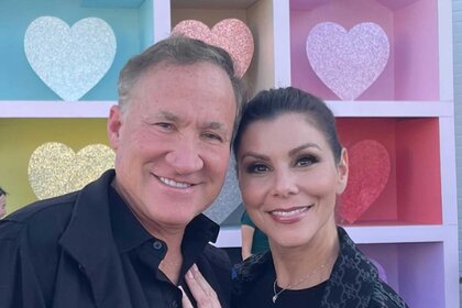 Heather Dubrow and Terry Dubrow pose together in front of a display with colorful, sparkly, hearts.