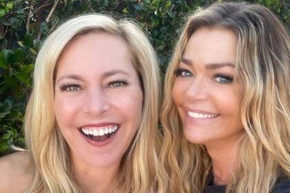 Sutton Stracke and Denise Richards smiling and laughing together while posing for a selfie in front of foliage.