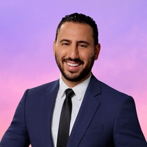 Josh Altman wearing a blue blazer in front of a purple and pink background.