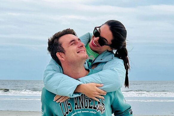 James Kennedy and Ally Lewber smiling and embracing each other at the beach on an overcast day.