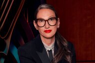 Jenna Lyons sitting in a black blazer and jeans in front of artwork and a wooden wall.