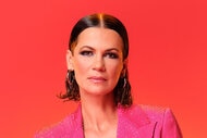 Julia Lemigova posing in a crystal embellished, hot pink, suit set in front of a red and orange background.