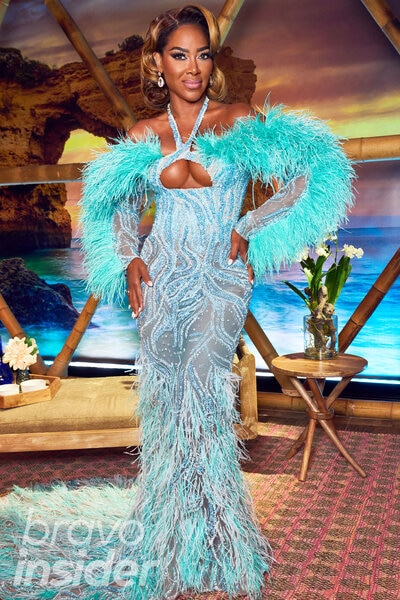 Kenya Moore in a teal feathered and crystal embellished fitted gown in front of a portugal inspired set.