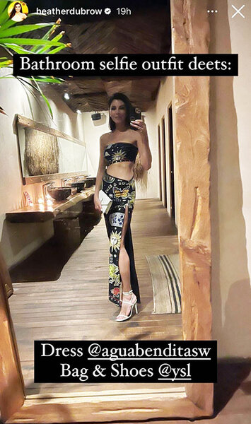 Heather Dubrow takes a selfie in front of a large bathroom mirror in a black patterned set. Overlaid text: “Bathroom selfie outfit deets: Dress @auguabenditasw Bag & Shoes @ysl”