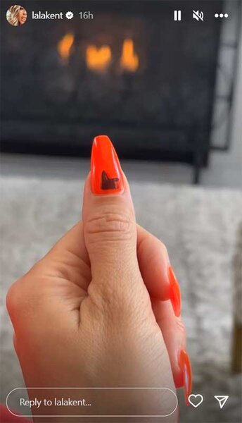 Lala Kent shows her orange nails in front of a fireplace.