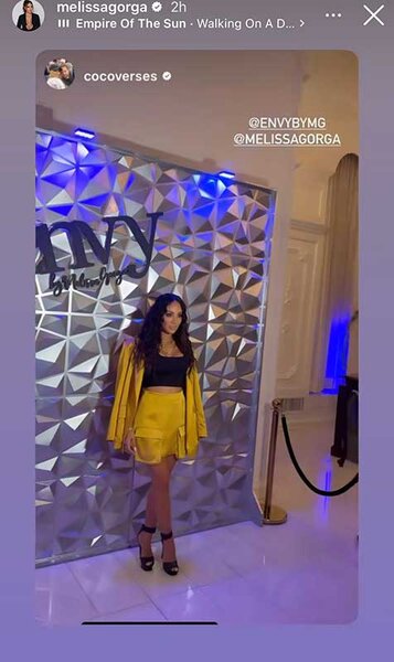 Melissa Gorga posing at her Envy fashion show in front of a textured wall wearing a yellow blazer and short set.