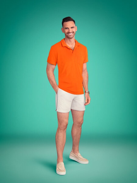 Full length of Carl Radke wearing an orange polo and light shorts in front of a green backdrop