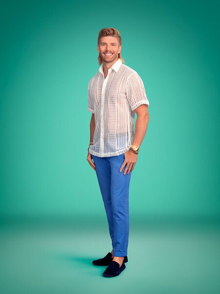 Full length of Kyle Cooke wearing a sheer white top and blue pants in front of a green backdrop
