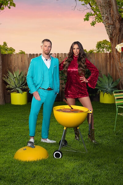 Kristen Doute wearing a red sequin dress and Luke Broderick wearing a blue suit in a backyard with a barbeque