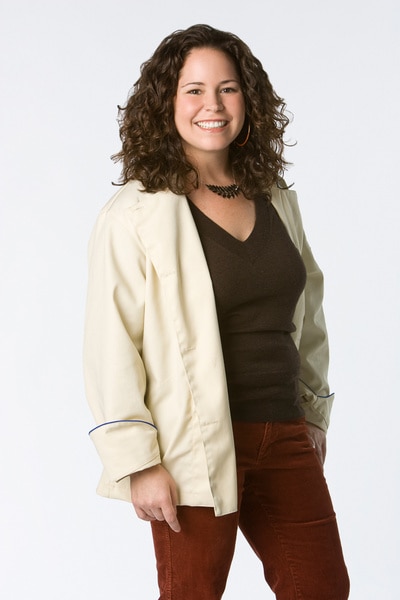 Stephanie Izard wearing her chef's jacket, red pants, and black tee in front of a white backdrop