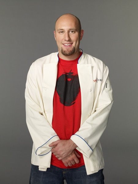 Hosea Rosenberg wearing his chef's jacket and red tee shirt in front of a grey backdrop
