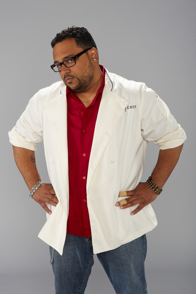 Kevin Sbraga wearing his chef's jacket and jeans in front of a grey backdrop.