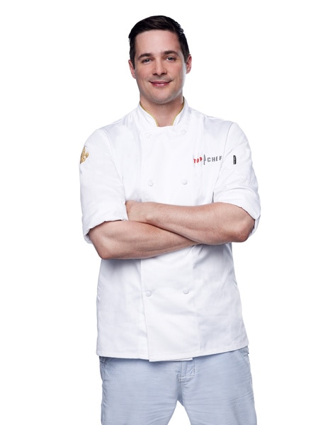 Nick Elmi wearing his chef's jacket and grey slacks in front of a white backdrop