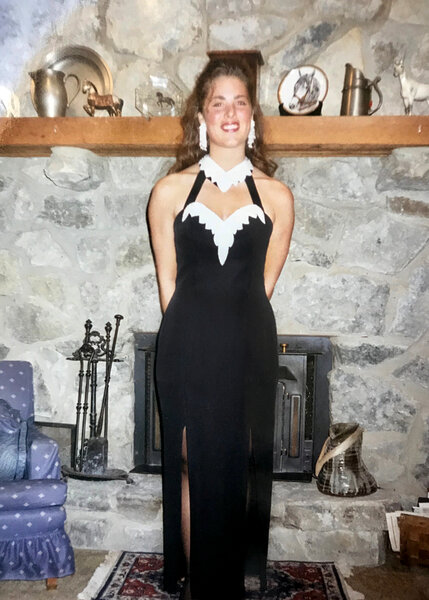 Emily as a teenager wearing a black gown with white embellishments in front of a fireplace.