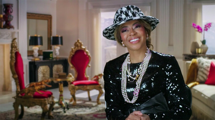 Mary Cosby smiling in a black-and-white outfit, a black-and-white hat, and long strands of pearls with a living room in the background.