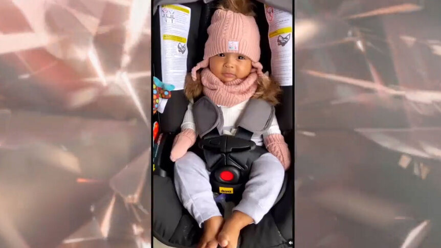 Mecca in a pink beanie hat and outfit strapped into a baby seat.
