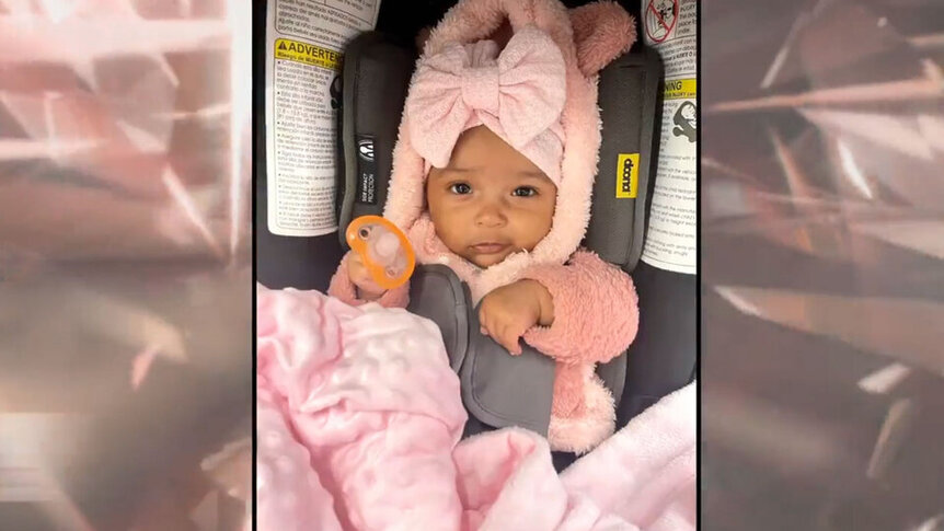 Mecca in a pink bow and outfit strapped into a baby seat.