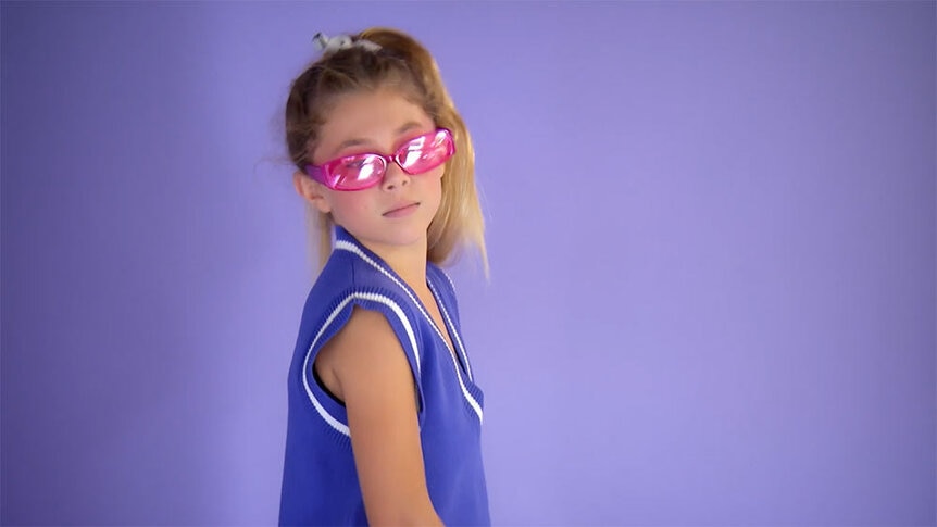 Annabelle modeling in pink sunglasses and a purple vest in front of a purple backdrop.