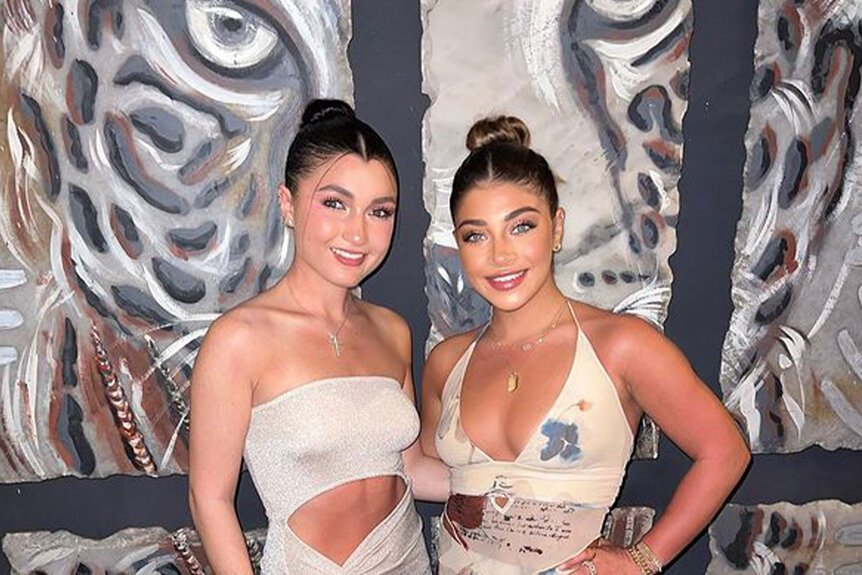 Gia and Gabriella pose together wearing updo's and neutral colored dresses in front of a tiger painting.