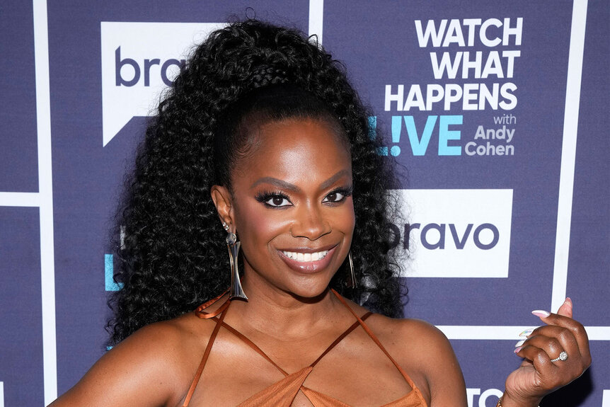 Kandi in a brown fitted minidress and a curly updo in front of the WWHL step and repeat.
