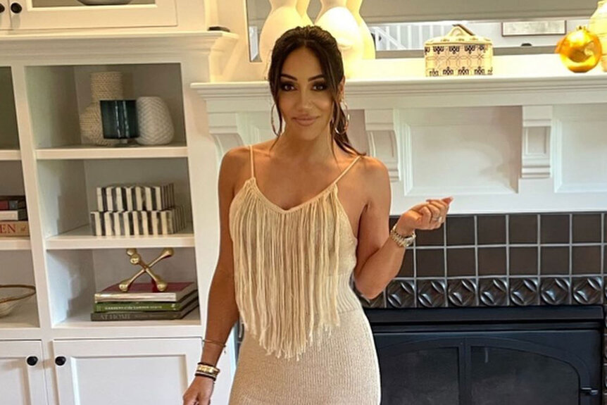 Melissa Gorga in a beige dress with fringe in front of a fireplace.