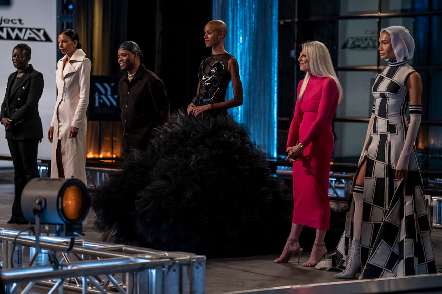 Project Runway finalists Brittany Allen, Bishme Cromartie, and Laurence Basse next to their designs on the Project Runway stage.
